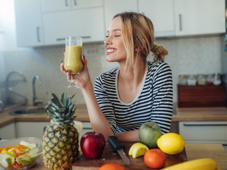 woman with yellow smoothie in the kitchen. In front of her are different vegetables and fruits on the countertop