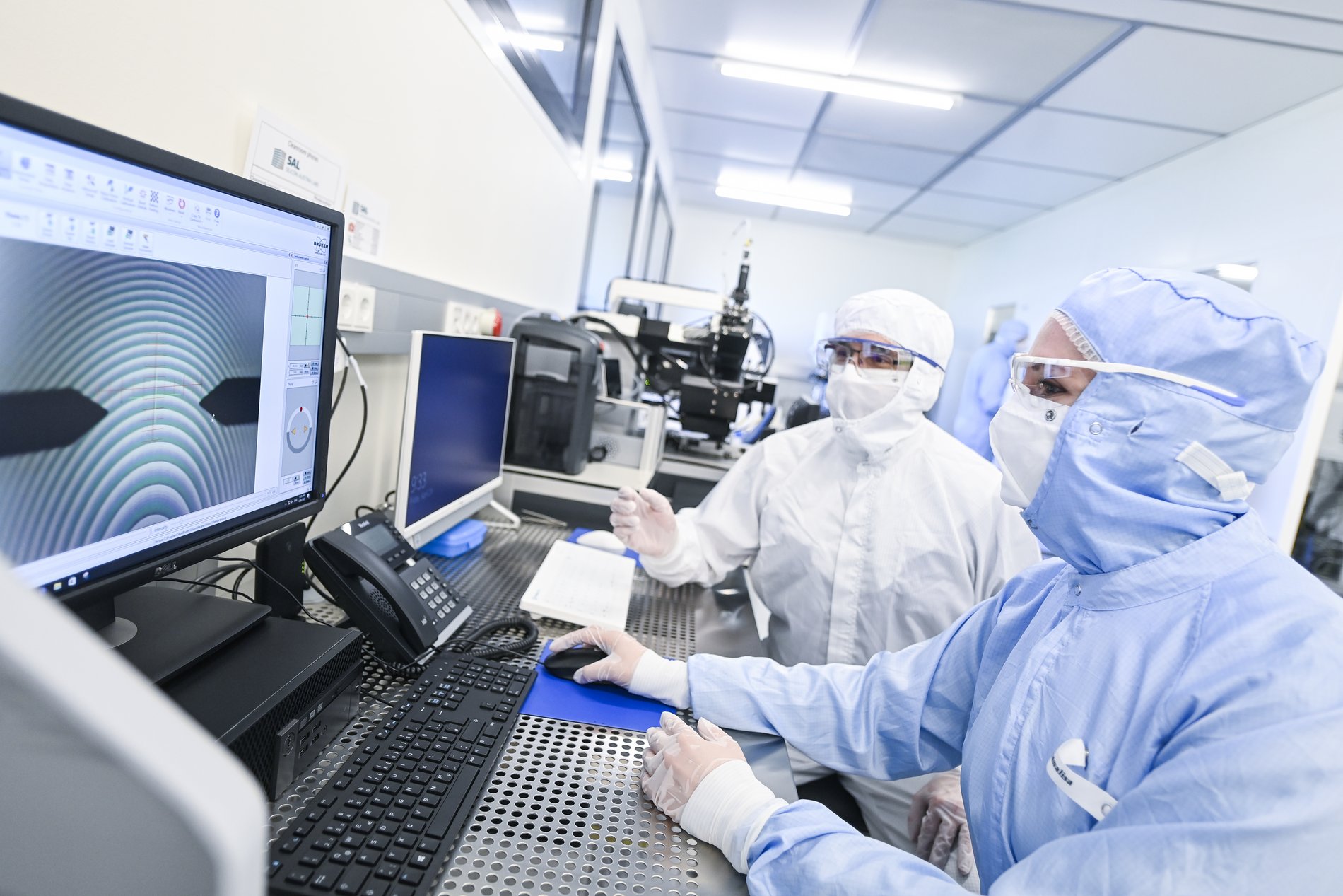 two researchers in protective clothing sit in front of monitors in a cleanroom