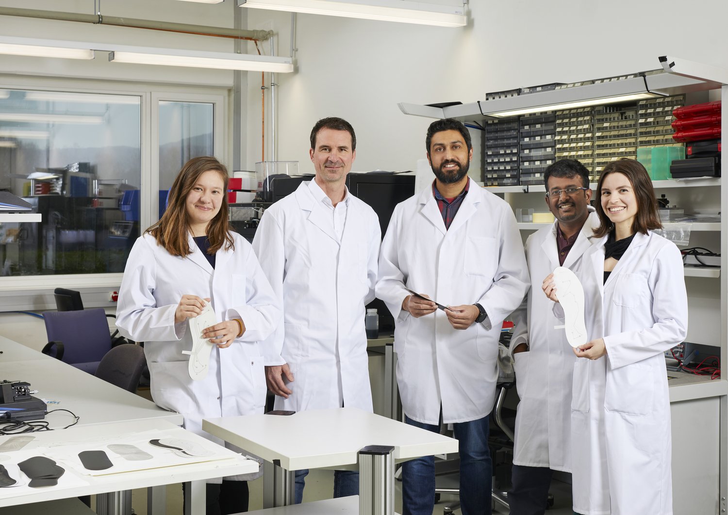 5 researchers in white lab coats, two are holding prototypes of the SOLES shoe inlay in their hands. 