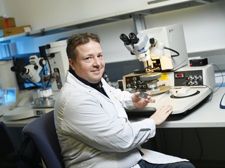 Ein Forscher sitzt vor einem Mikroskop und schaut in die Kamera