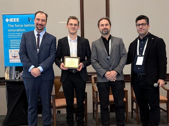 Four men in suits. In the middle is Alexander, holding the best paper award. Next to him Hans-Peter Bernhard, Head of Wireless Communications. To their left and right two people from the WF-IoT Conference. 