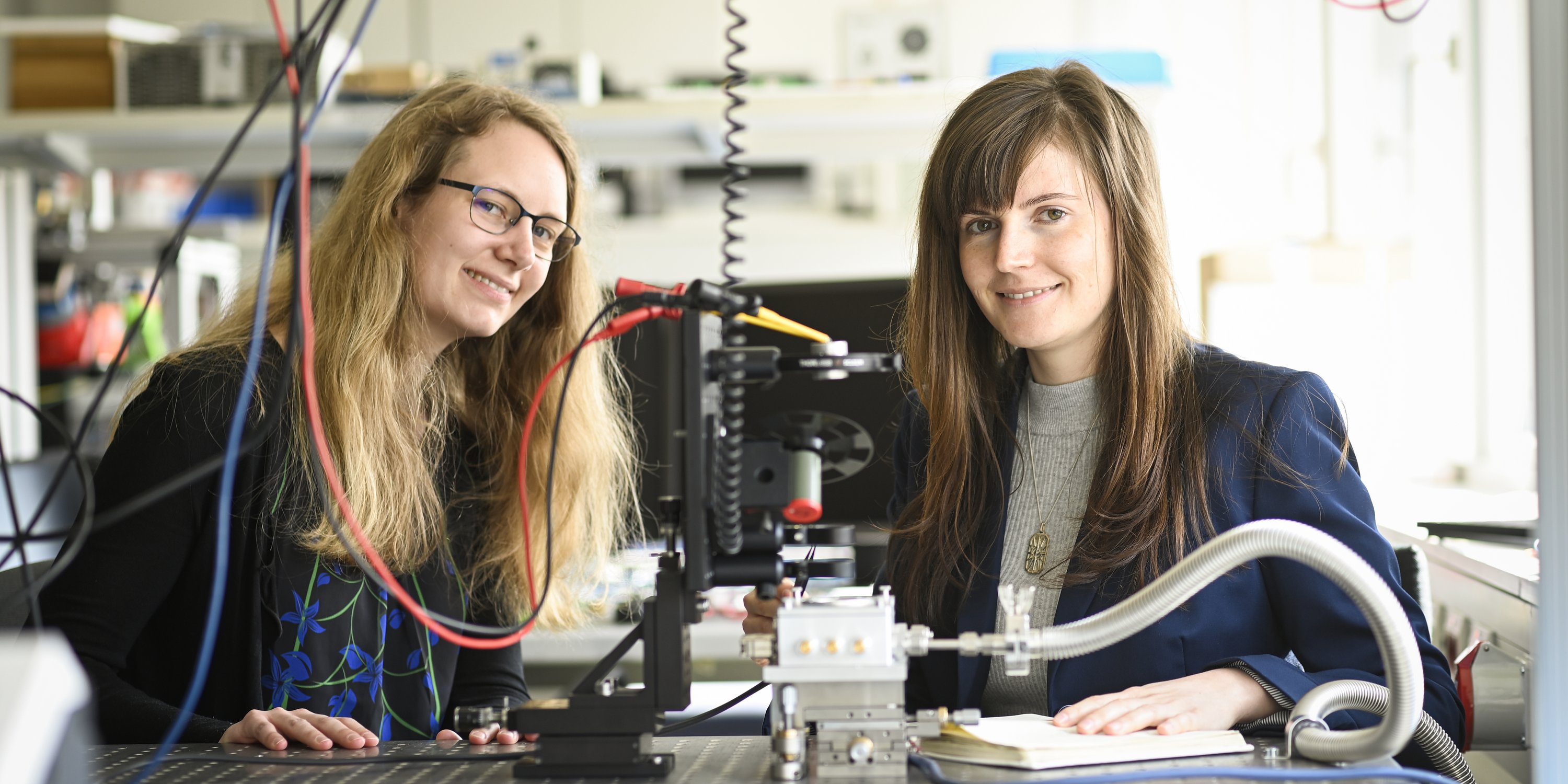 zwei Forscherinnen sitzen im Labor und lachen in die Kamera