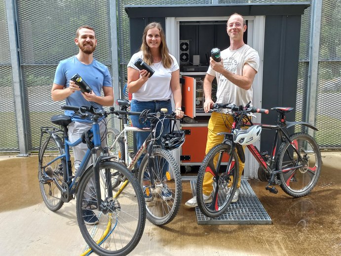 3 employees holding bike and battery to power the bike electrically; charging station in the background