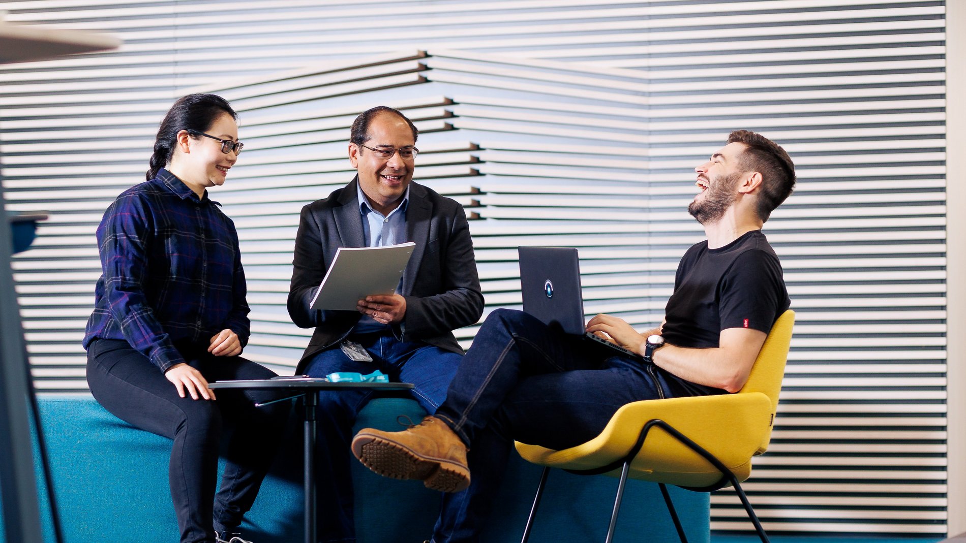 drei Personen sitzen mit ihren Laptops auf bunten Büromöbeln und lachen